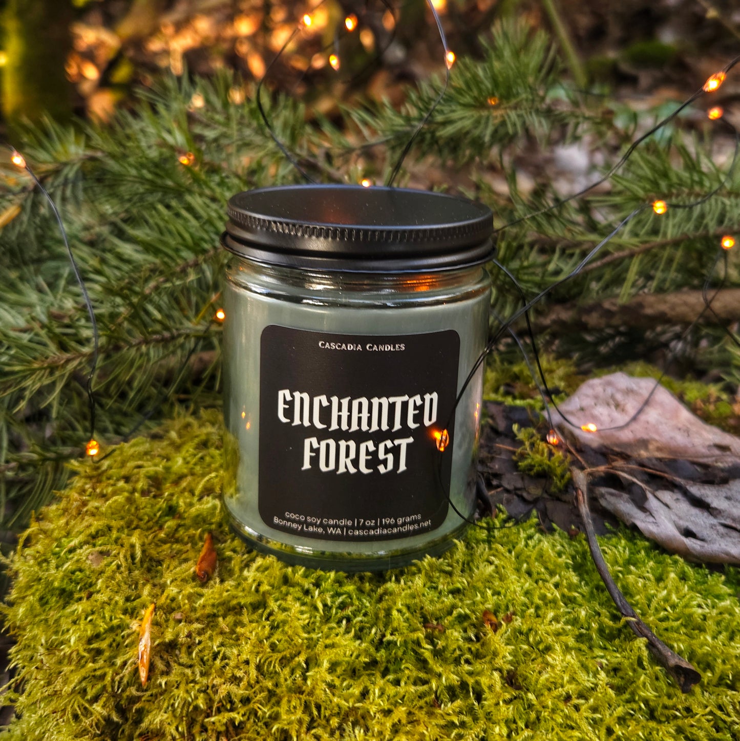Candle labeled 'Enchanted Forest' on a mossy surface with greenery and lights in the background