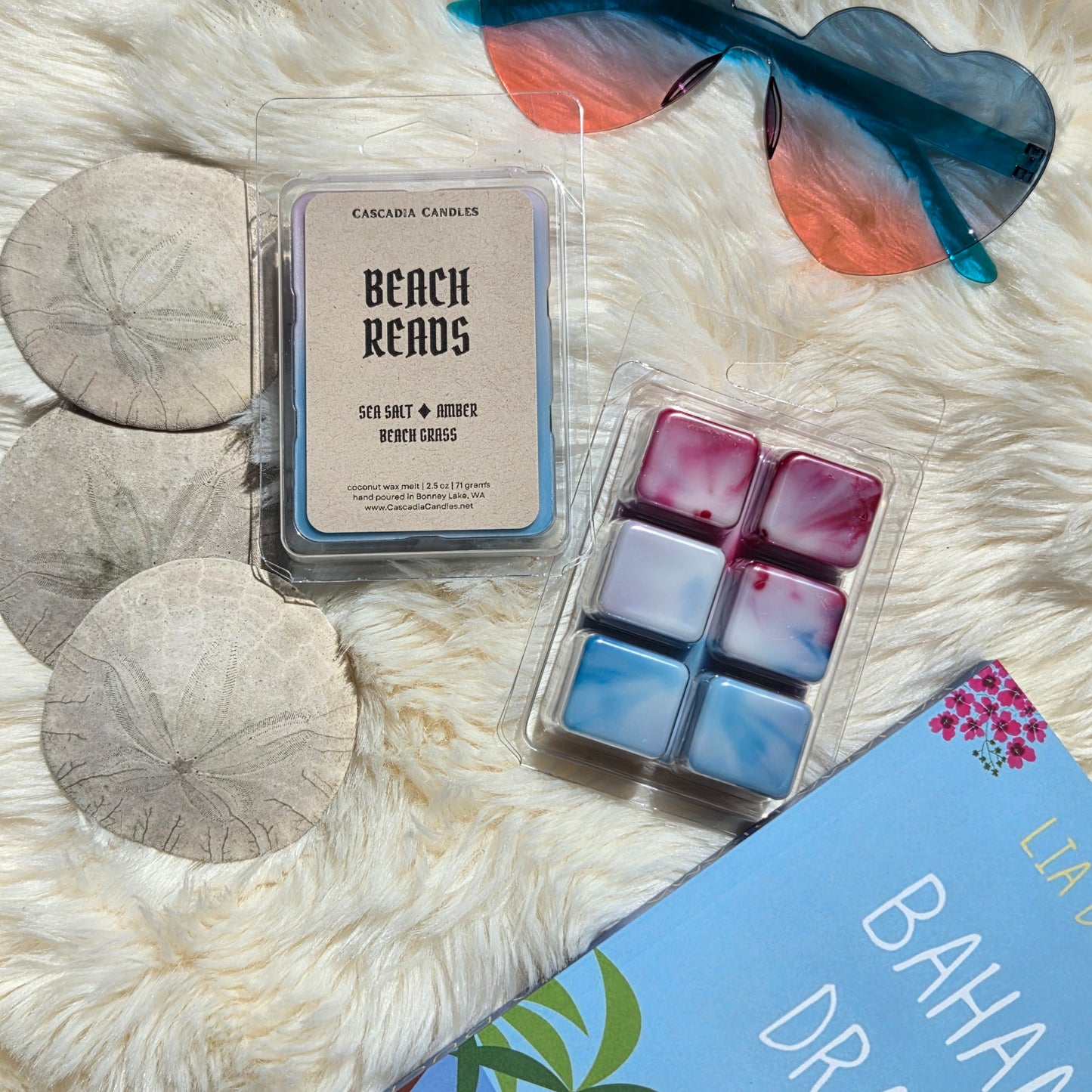 Packaged wax melts with a label on a fluffy white surface with sunglasses and sand dollars.