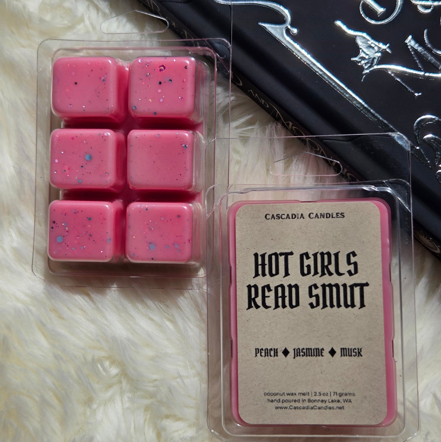 Two packaged wax melts from Cascadia Candles on a white faux fur surface: six pink wax cubes in a clear clamshell on the left, and a single pink-bordered clamshell labeled 'HOT GIRLS READ SMUT' with scent notes 'PEACH + JASMINE + MUSK' on the right. A black patterned book is partly visible behind the products.