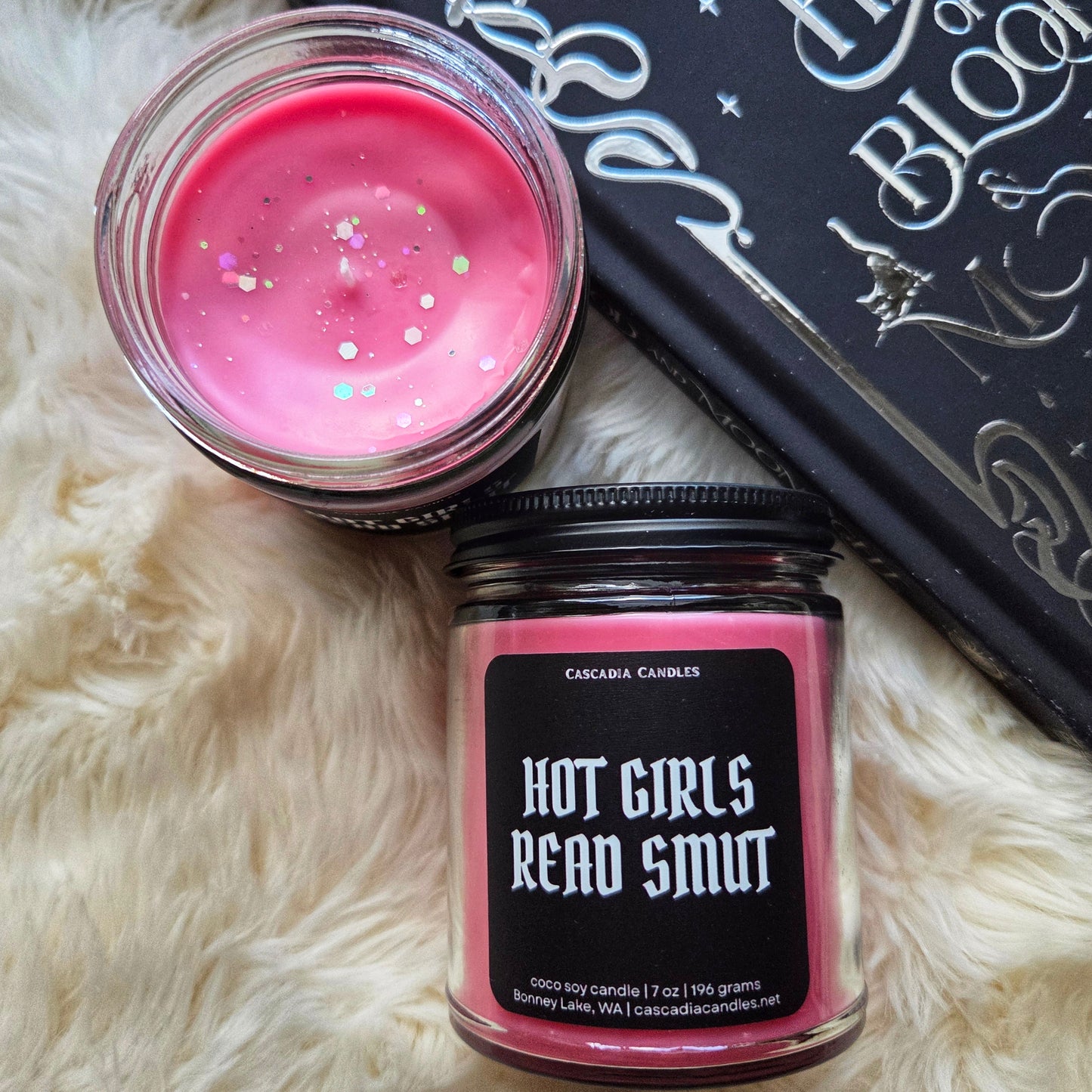 'Hot Girls Read Smut' candle next to black and silver book on white fluffy surface.