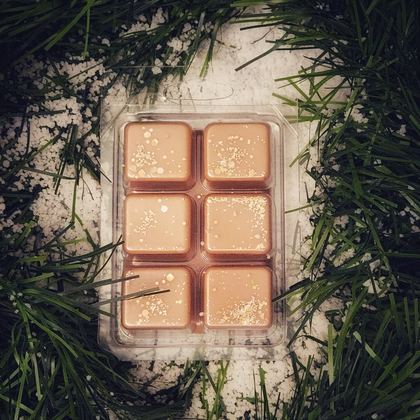 Pack of six square wax melts with glittery texture on a bed of green garland and snow.