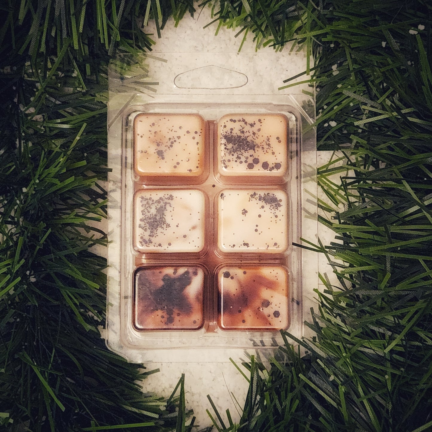 Pack of six square wax melts in a clear plastic packaging on a bed of green garland and snow.