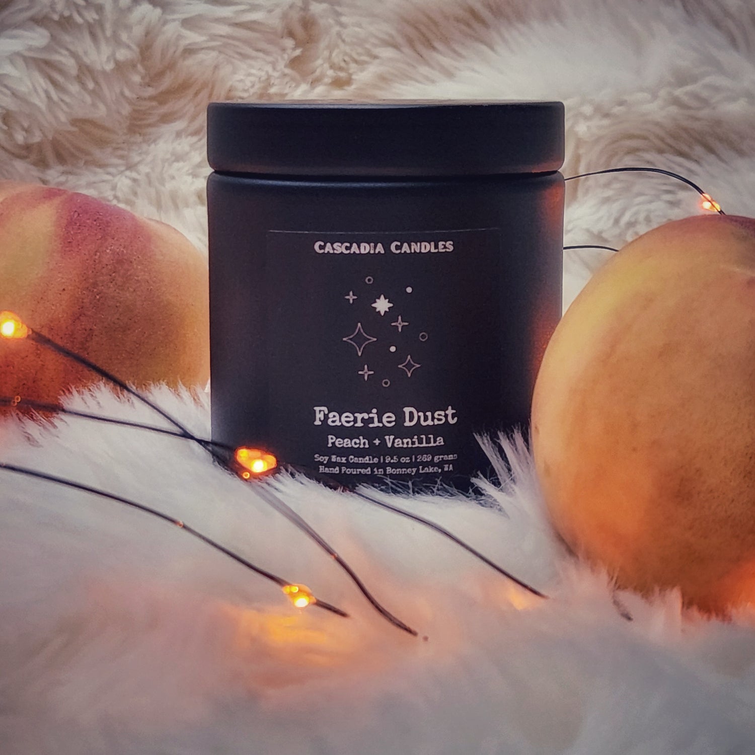 A black jar candle labeled 'Faerie Dust' with a mix of decorative string lights and peaches in the background, on a furry white surface.