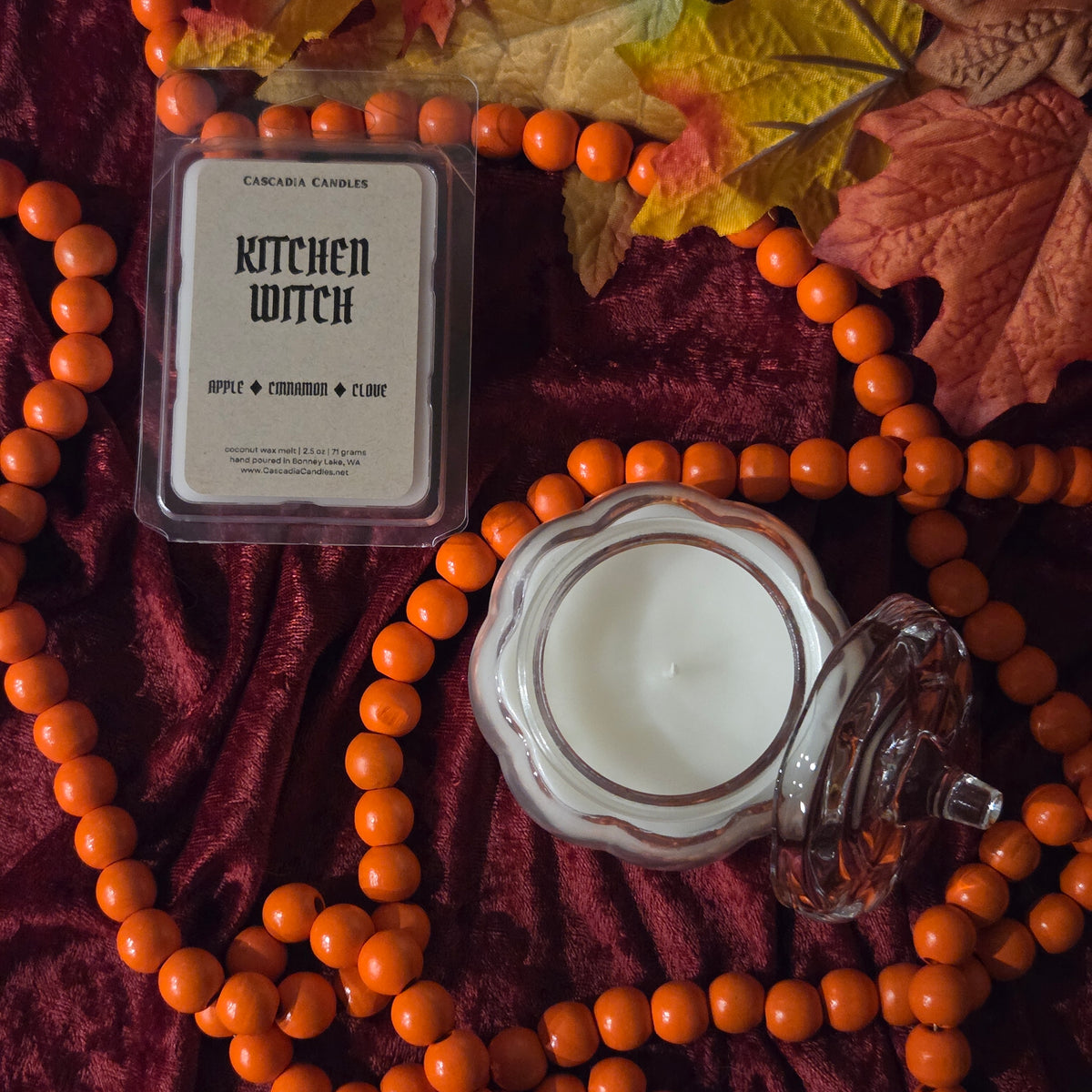 A styled flat-lay of a scented candle product on a deep burgundy velvet fabric, surrounded by orange beads and autumn leaves. The product includes a clear plastic clamshell package labeled 'KITCHEN WITCH' by Cascadia Candles, and an open glass pumpkin-shaped jar containing white wax.
