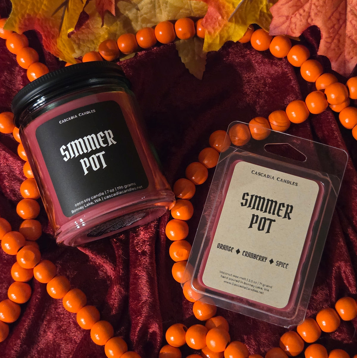 A Simmer Pot candle and a Simmer Pot wax melt side-by-side on a red velour background with orange garland and fall leaf decor.