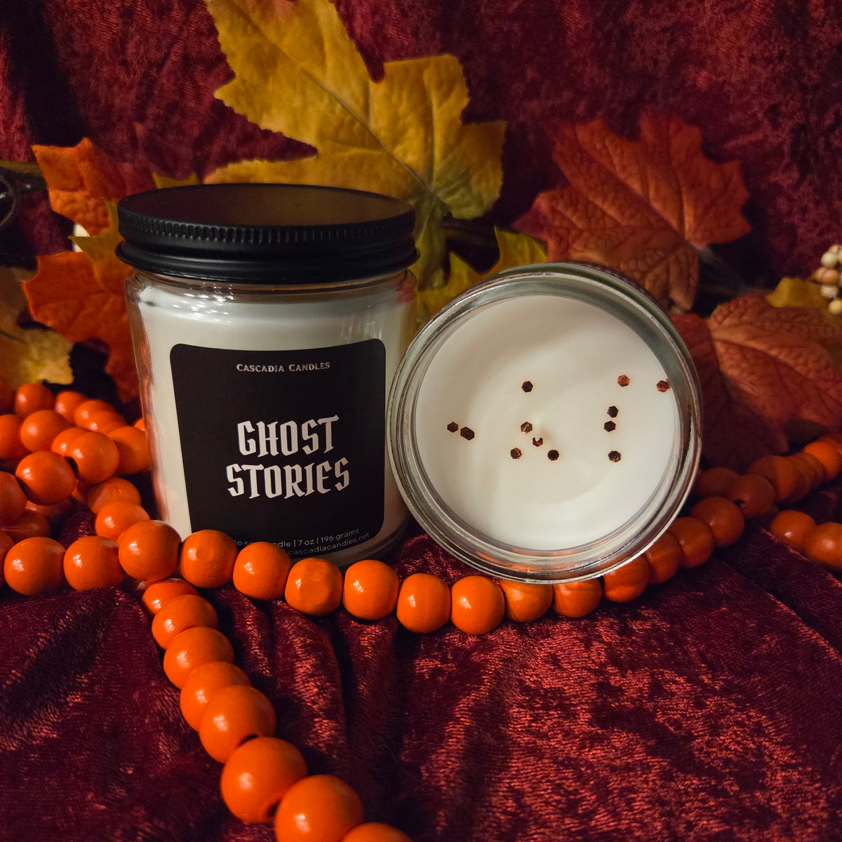 A jar candle labeled 'GHOST STORIES' by Cascadia Candles, shown with the lid off to reveal white wax and copper eco-friendly glitter on top, styled on a dark red velvet fabric with orange beads and autumn leaves.