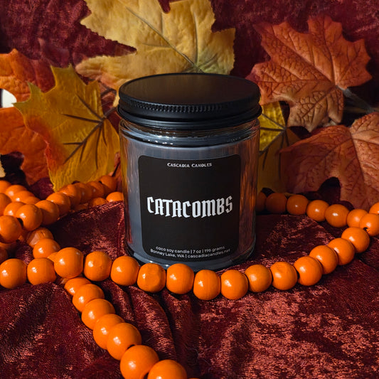 A glass jar candle with a black lid and a black label reading 'catacombs' sits on dark red velvet fabric, surrounded by orange beads and autumn leaves.