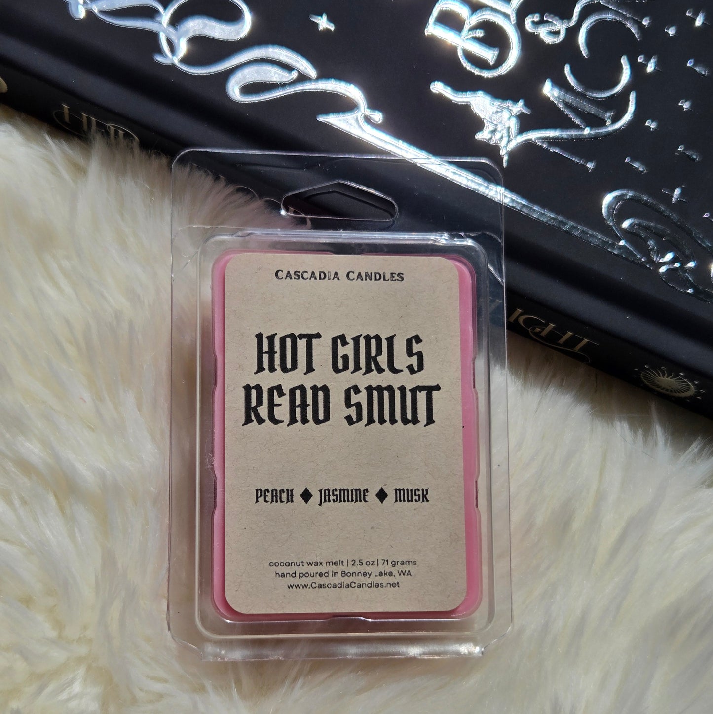 A packaged wax melt from Cascadia Candles labeled 'Hot Girls Read Smut' in peach, jasmine, and musk scents, placed on a white faux fur surface with a black patterned book in the background.