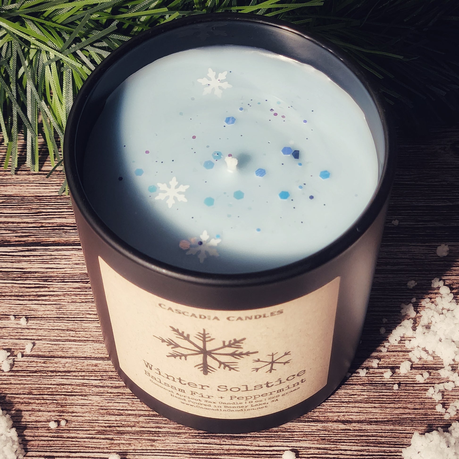 Candle with snowflake decorations in a decorative container on a wooden surface with greenery.