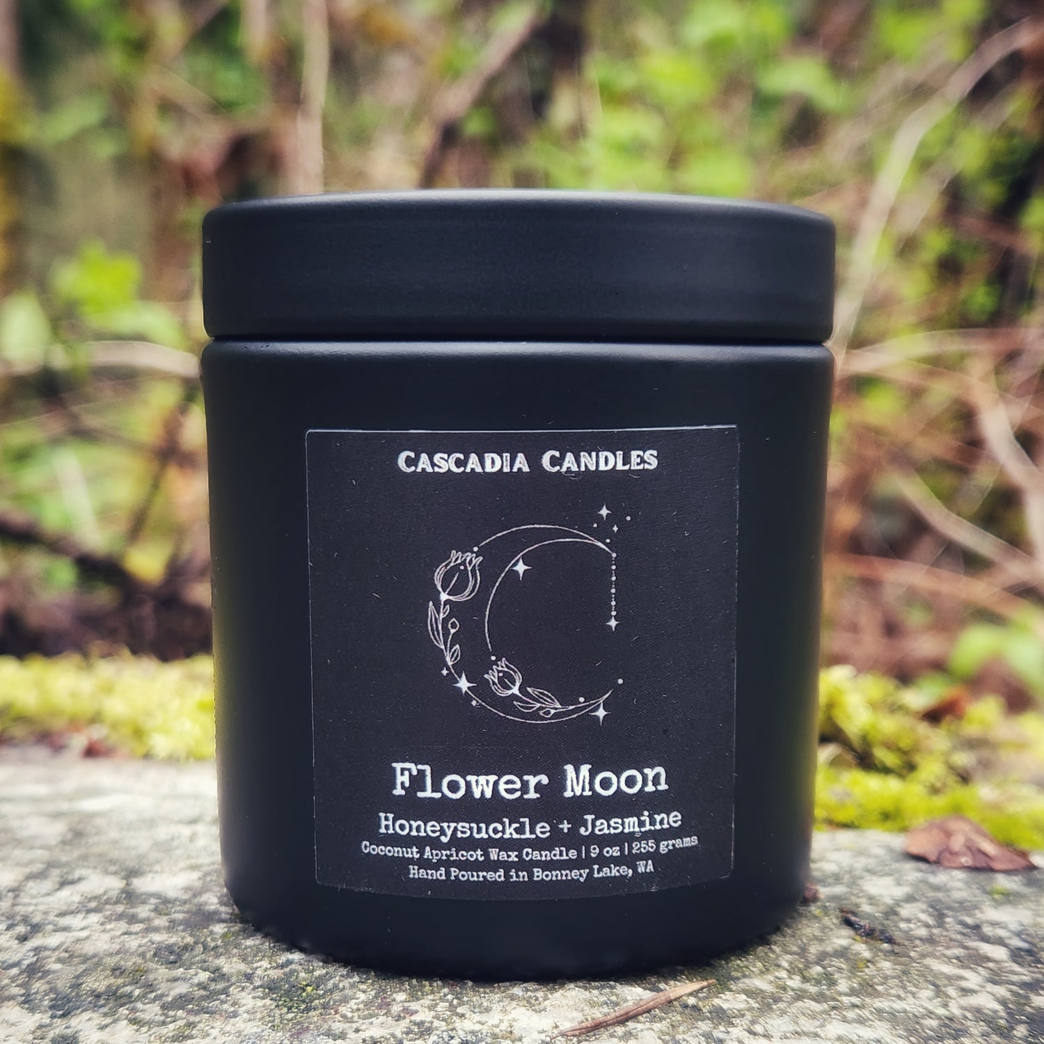 A black tin candle with a label reading 'Cascadia Candles, Flower Moon, Honeysuckle & Jasmine'. The candle is set against a blurred background with natural elements.