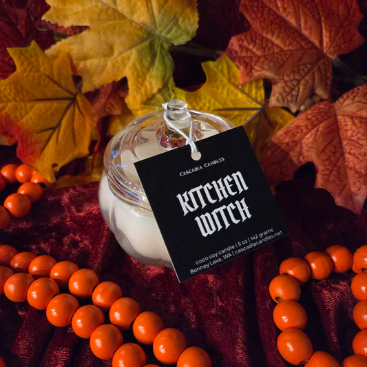A glass jar candle with white wax sits on dark red velvet fabric, surrounded by orange beads and autumn leaves. A black hanging tag reads 'KITCHEN WITCH' and CASCADIA CANDLES.