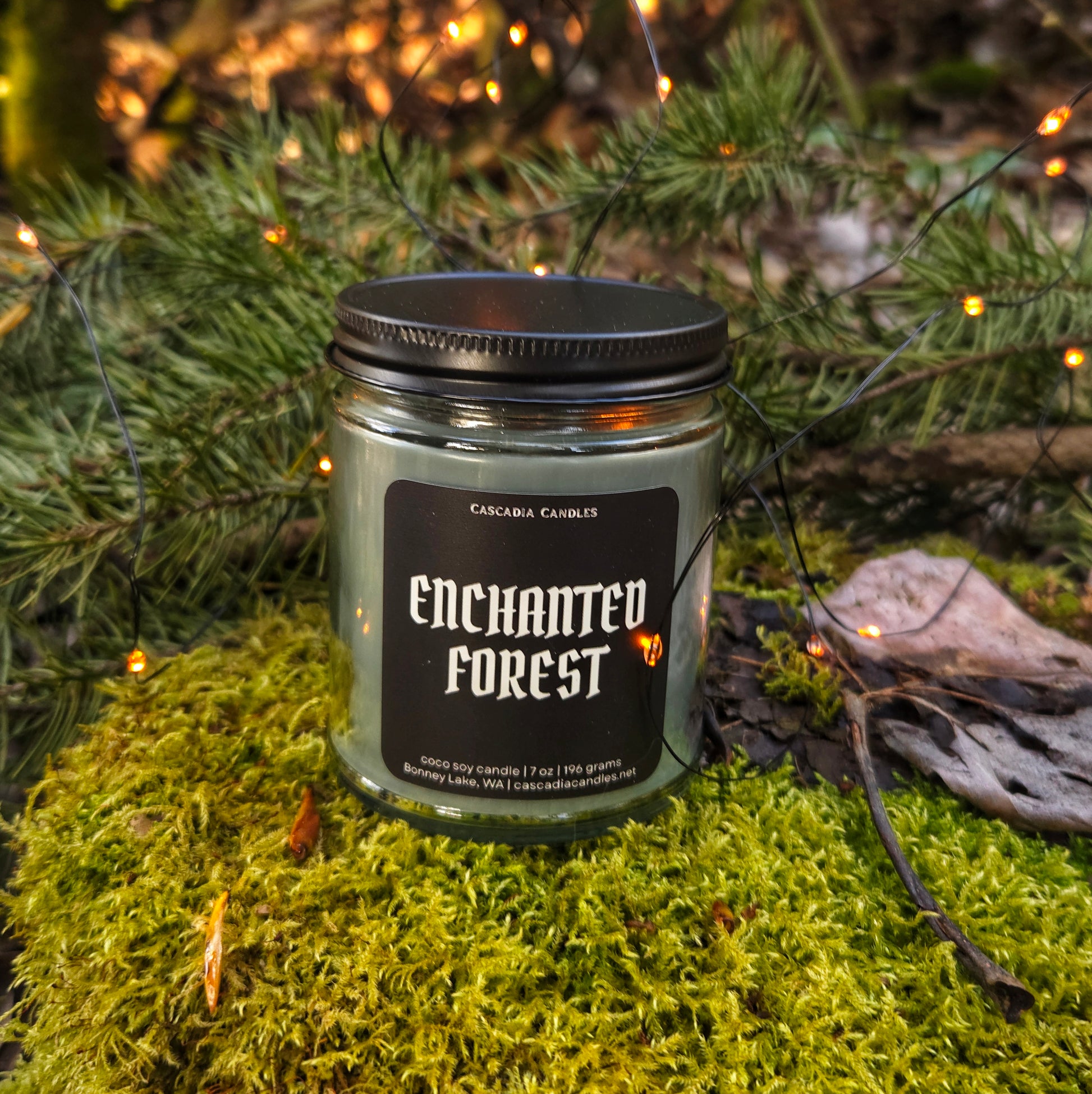 Candle labeled 'Enchanted Forest' on a mossy surface with greenery and lights in the background