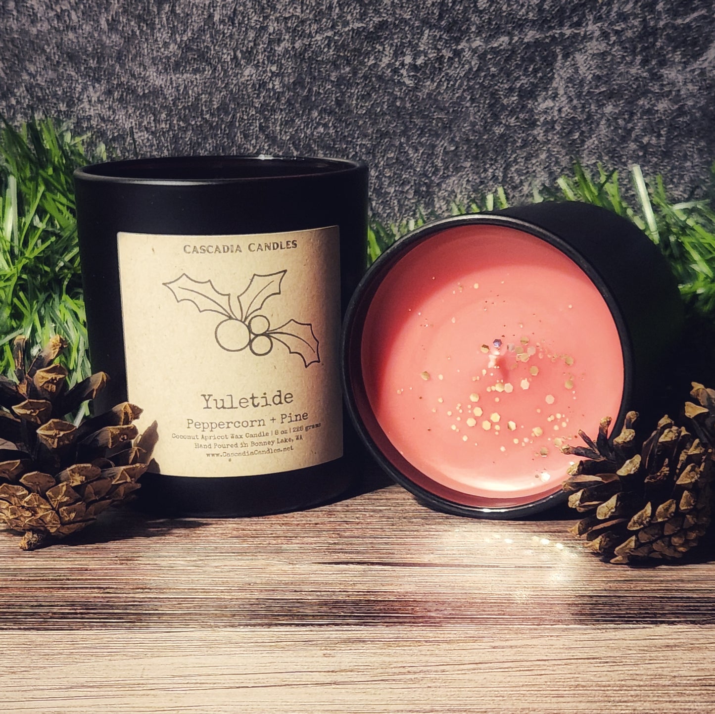 Candle with a visible label on a wooden surface with pinecones and grass in the background