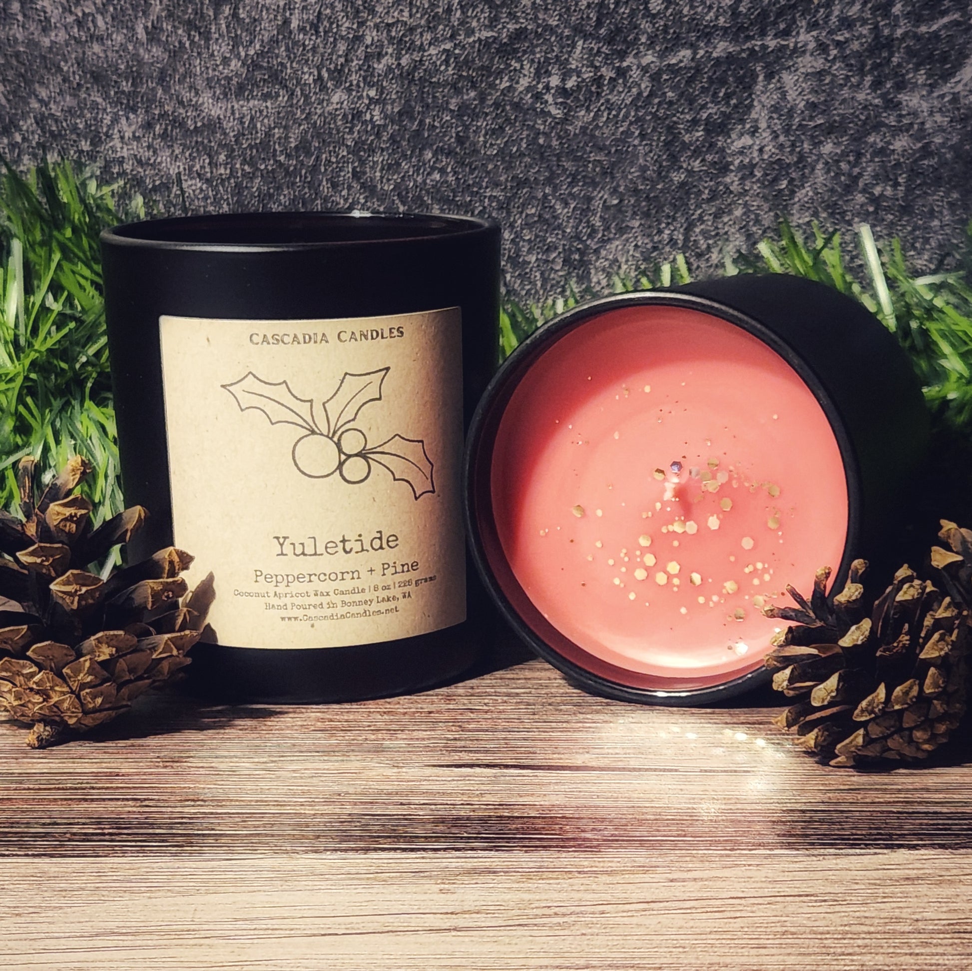 Candle with a visible label on a wooden surface with pinecones and grass in the background