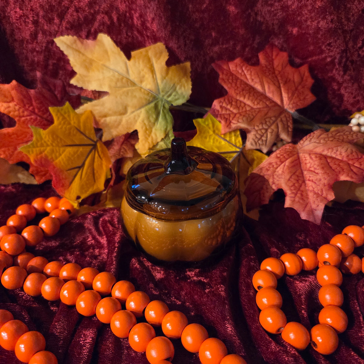 A small amber glass pumpkin-shaped jar with a lid, styled on a dark red velvet fabric with autumn leaves and orange bead garlands.