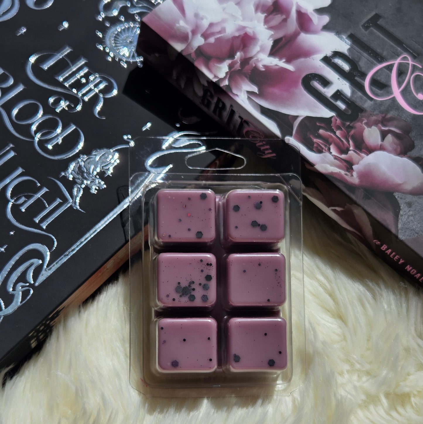 Six purple wax melts with black eco-friendly glitter in a clear plastic clamshell package, placed on a white faux fur surface next to a black book and a floral book cover.