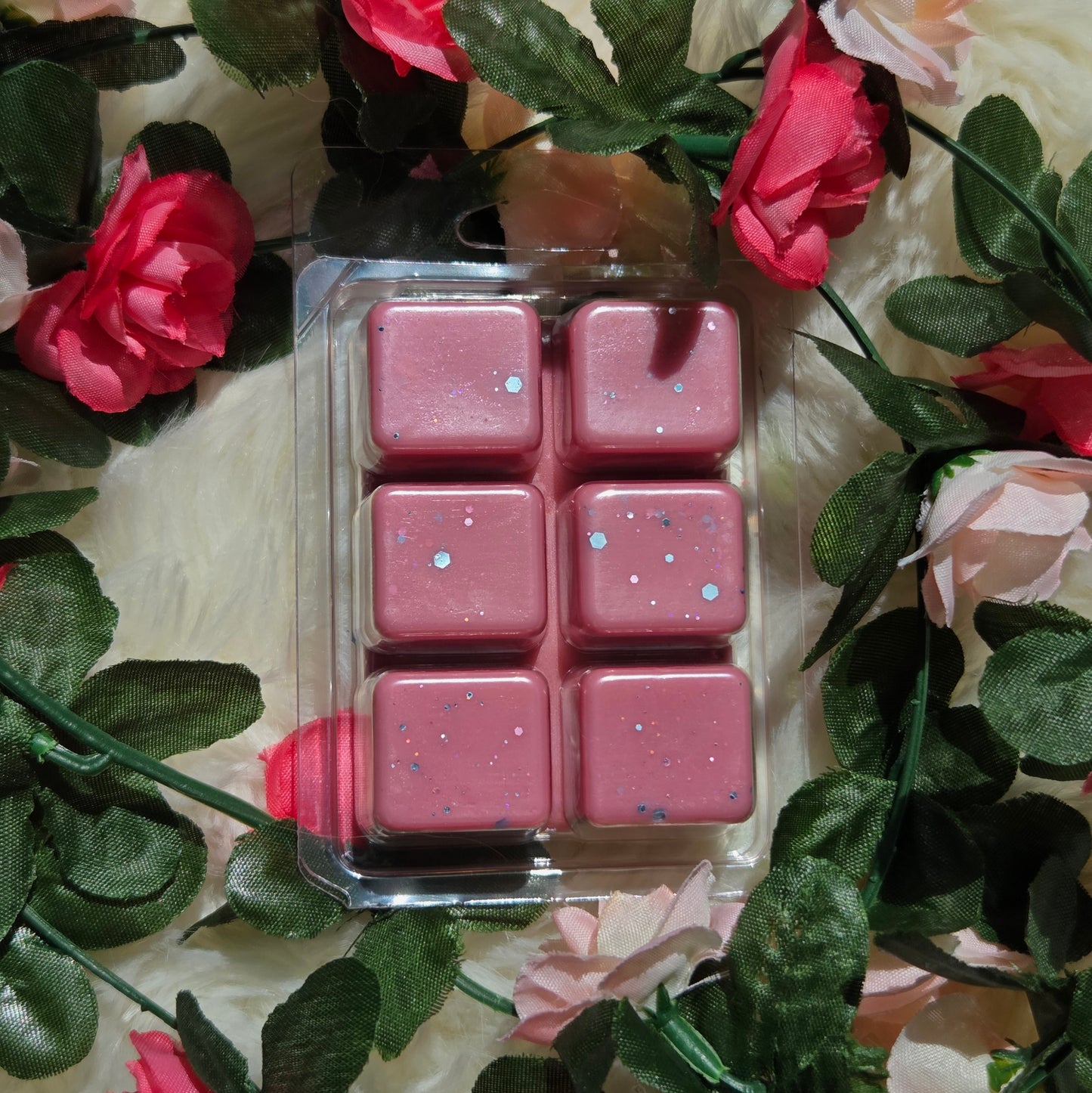 Six pink wax melts with eco-friendly glitter in a clear plastic clamshell package, surrounded by artificial pink roses and green leaves on a white faux fur background.