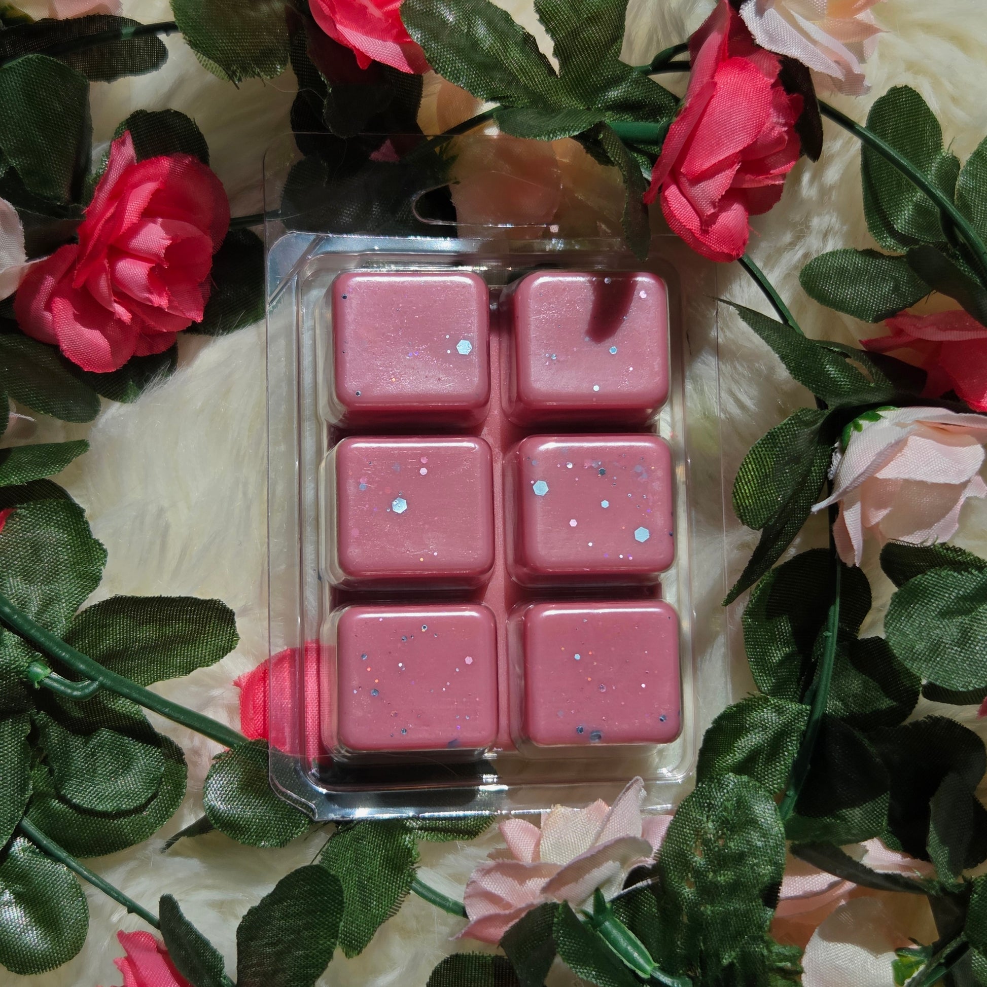 Six pink wax melts with eco-friendly glitter in a clear plastic clamshell package, surrounded by artificial pink roses and green leaves on a white faux fur background.