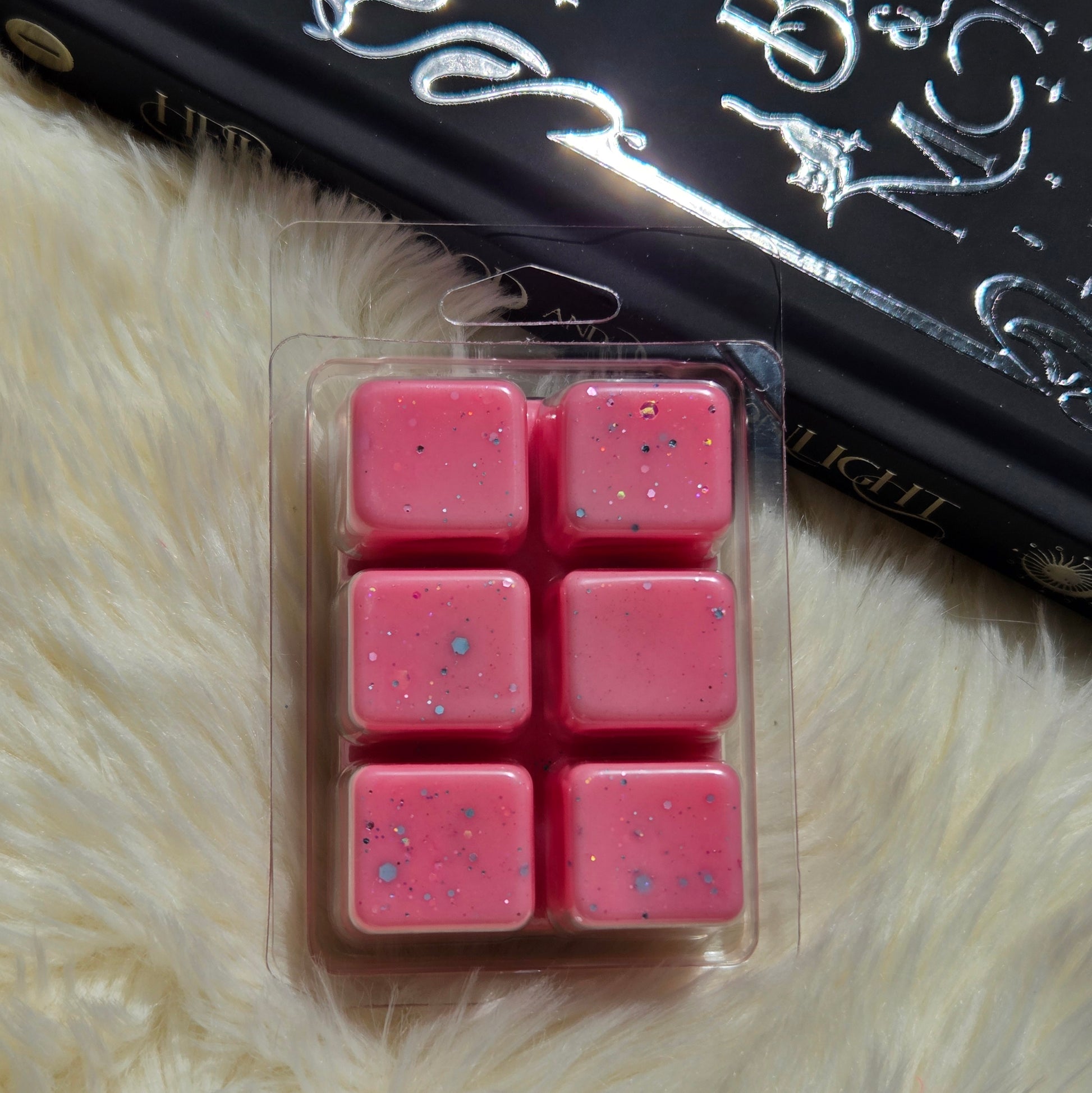 Six pink wax melts with blue and silver glitter flakes, packaged in a clear blister pack, placed on a white faux fur surface next to a black book.