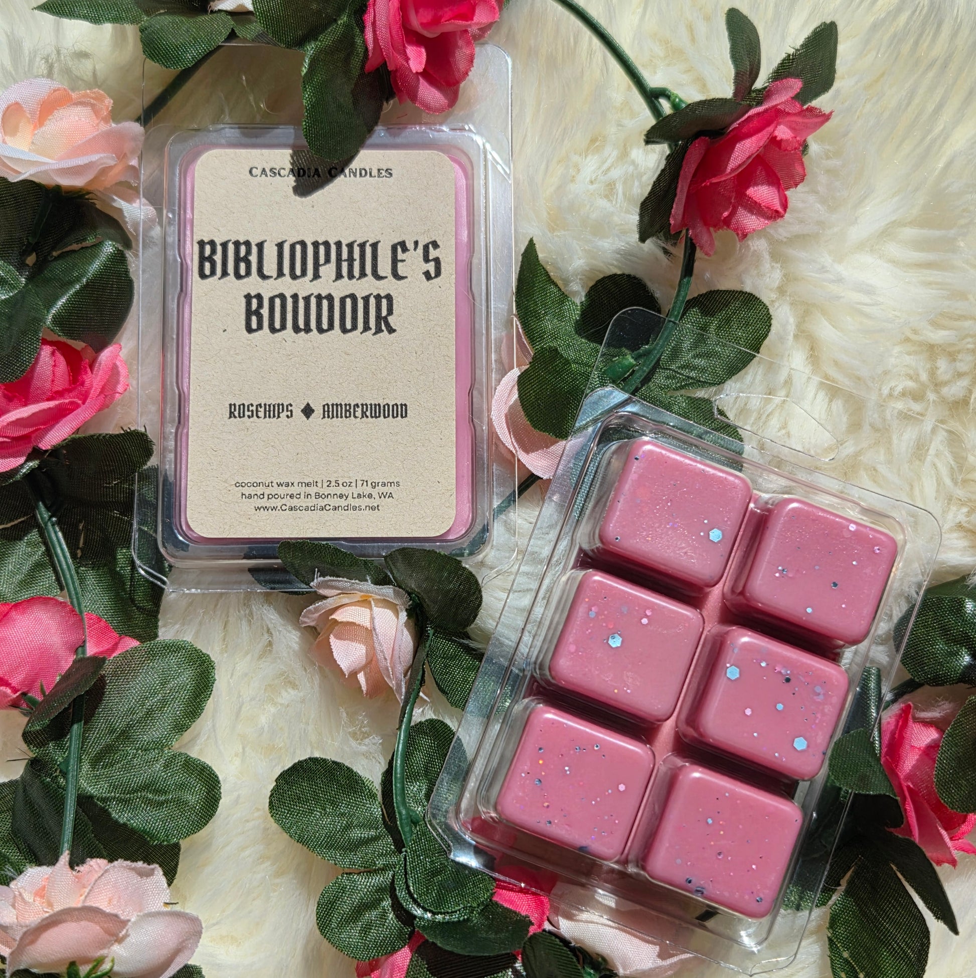 Two packaged wax melts from Cascadia Candles, one labeled 'Bibliophile's Boudoir' and the other showing six pink wax cubes with blue and silver eco-friendly glitter, styled with pink roses and green leaves on a white faux fur background.