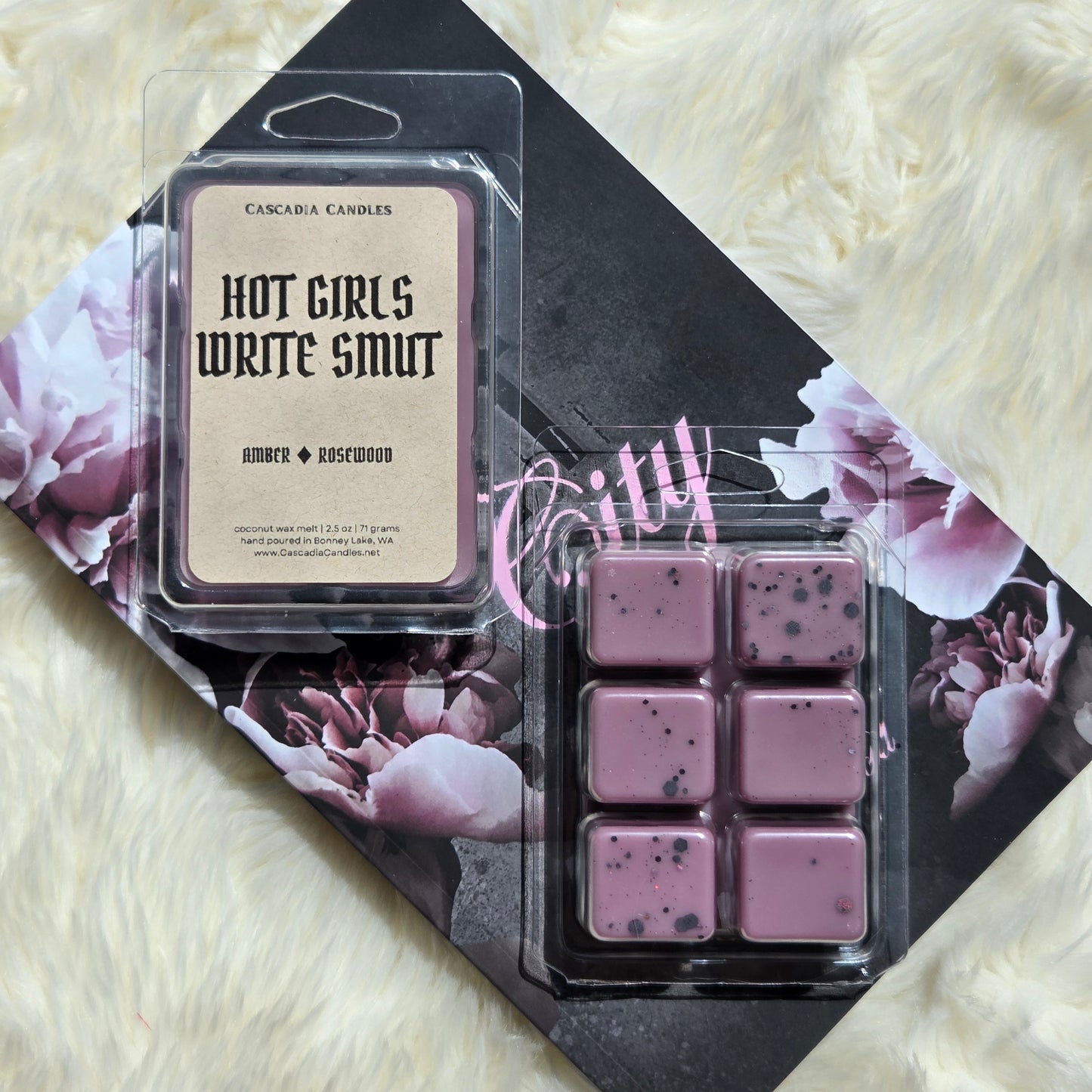 A packaged wax melt set from Cascadia Candles on a floral book cover, placed on a white faux-fur surface. The set includes a single clamshell wax melt labeled 'Hot Girls Write Smut' in an amber and rosewood scent, and a six-piece wax melt clamshell showing dusty mauve wax cubes with black holographic eco-friendly glitter.
