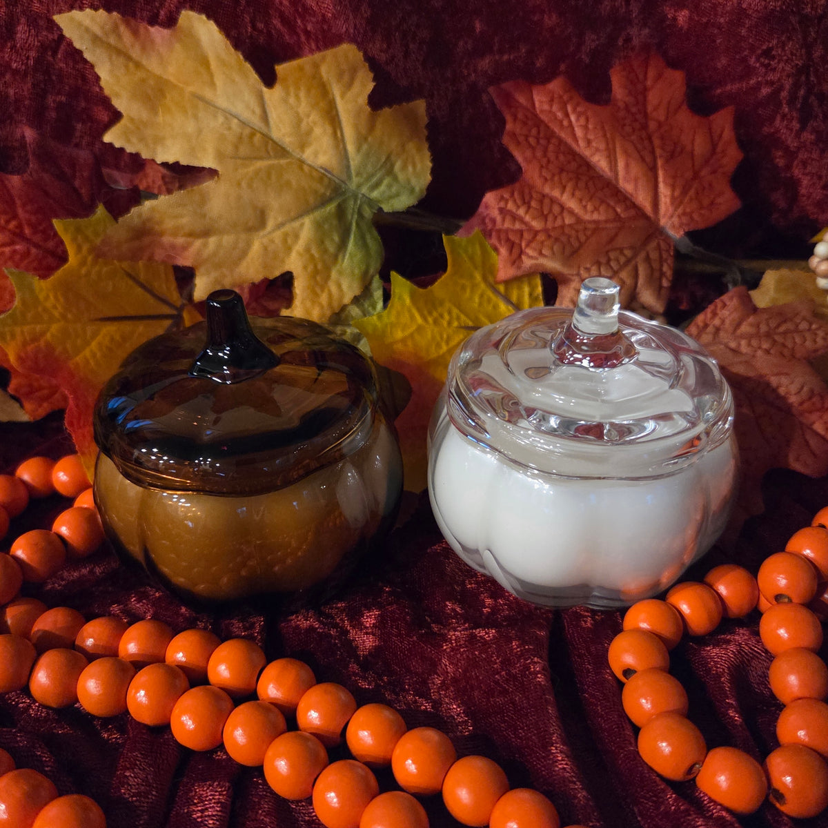 Two decorative glass pumpkin-shaped jars styled with autumn props, including orange beads, artificial leaves, and a dark red fabric backdrop. One jar is amber-brown and the other is clear glass containing white wax, both with matching pumpkin stem lids.