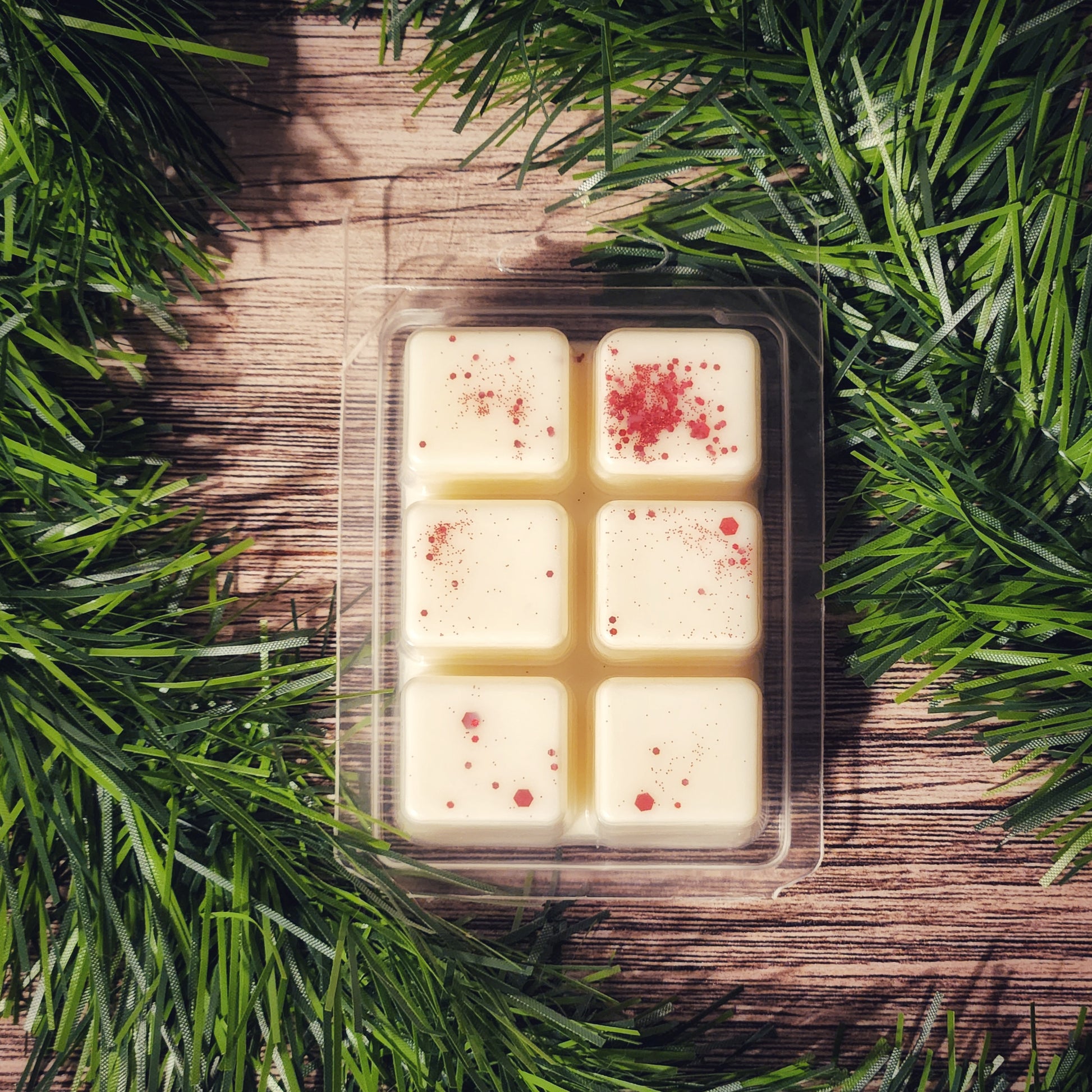 Pack of six white wax melts with red specks in a clear plastic packaging on a wooden surface surrounded by green garland.