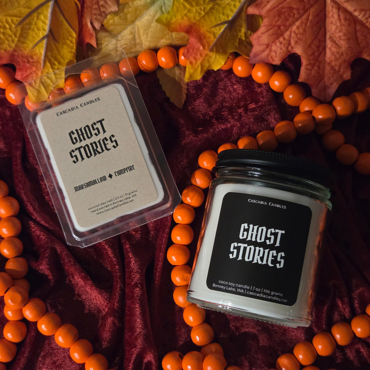 Two scented products from Cascadia Candles, one in a clear clamshell package and one in a glass jar with a black lid, both labeled 'GHOST STORIES,' arranged on dark burgundy fabric with orange beads and autumn leaves.