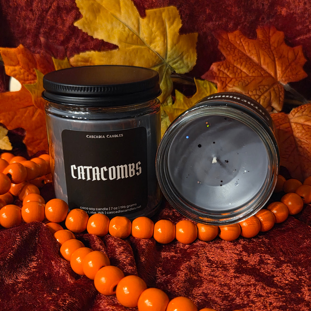 A jar candle labeled 'Catacombs' by Cascadia Candles, shown with its black lid beside the open glass jar. The candle is styled with orange beads and autumn leaves on a textured red fabric background.