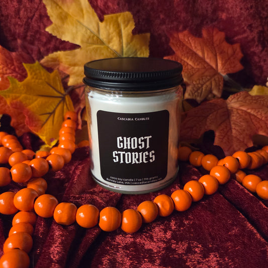 A jar candle labeled 'GHOST STORIES' by Cascadia Candles sits on a dark red velvet fabric surrounded by orange beaded garland and autumn leaves. The candle has white wax and a black lid.