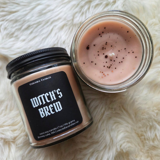 Two candles labeled 'Witch's Brew' on a fluffy white surface