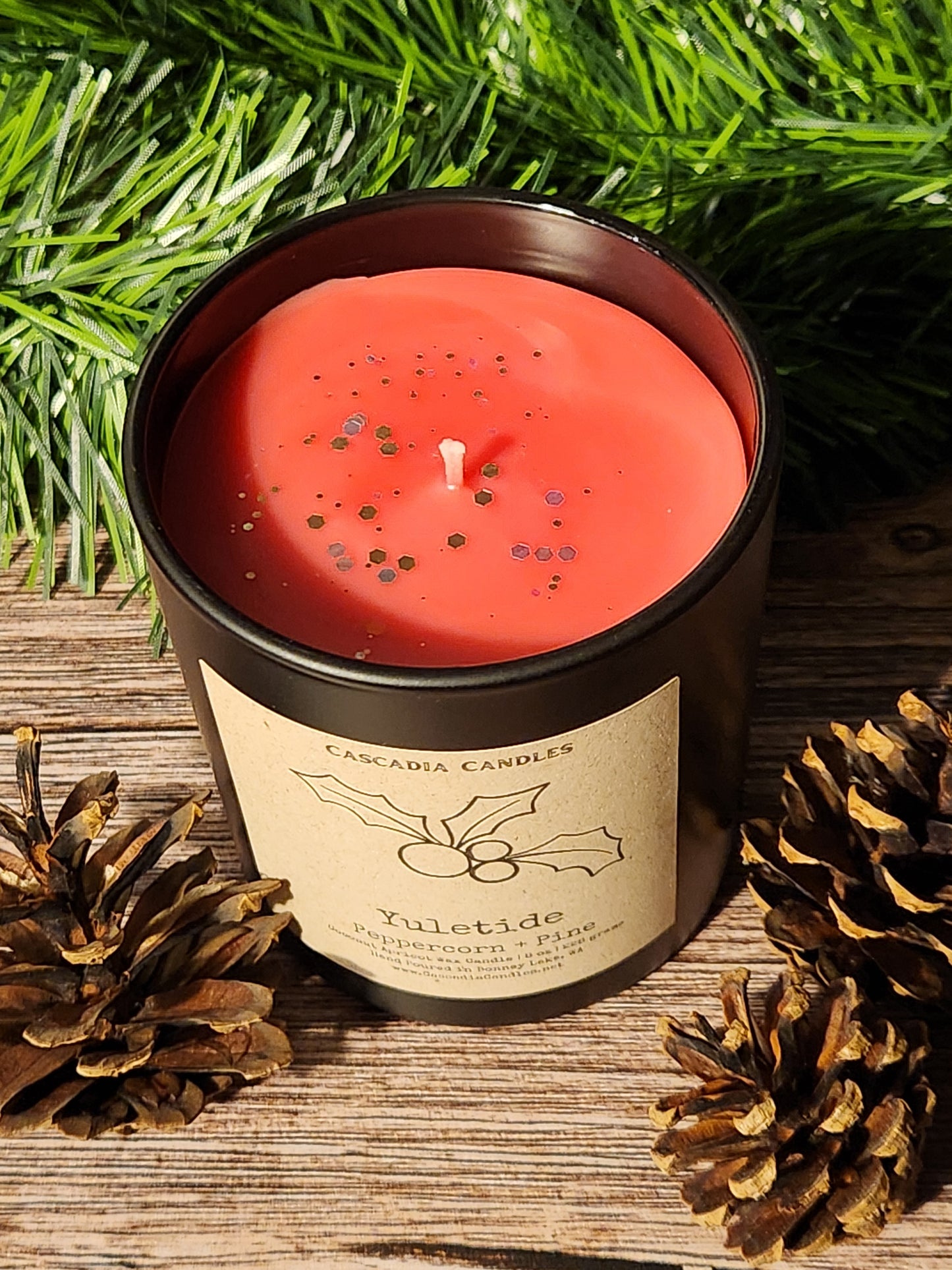 Red candle in a tin with a label, surrounded by pine branches and cones on a wooden surface.
