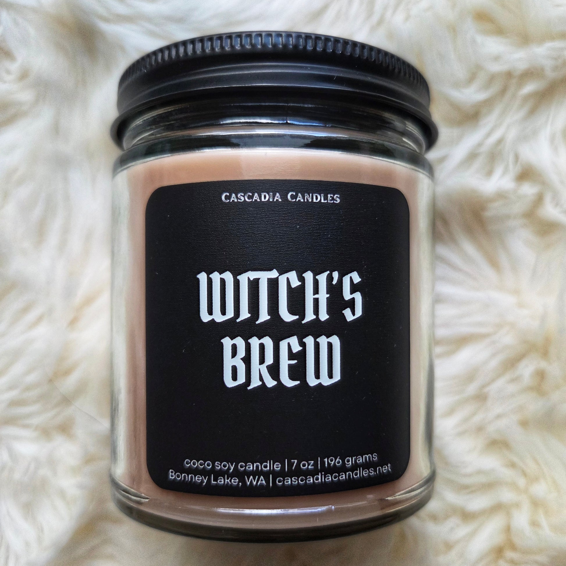 Candle jar labeled 'Witch's Brew' by Cascadia Candles on a white fluffy surface