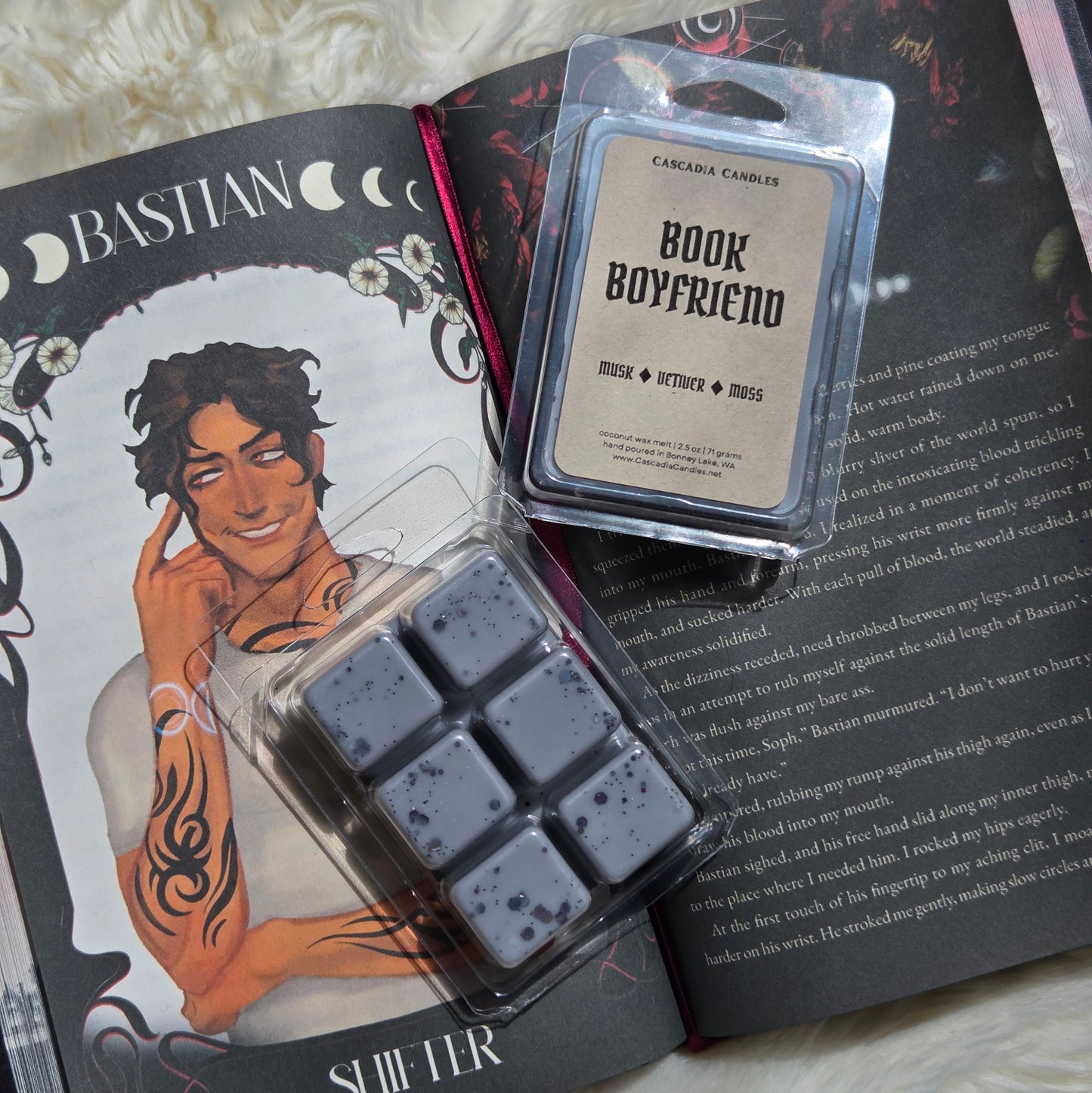 Two packaged wax melts are arranged on an open book. One package contains dark gray speckled wax cubes in a clear plastic clamshell, and the other is a clear blister pack with a Kraft label reading 'Book Boyfriend'.