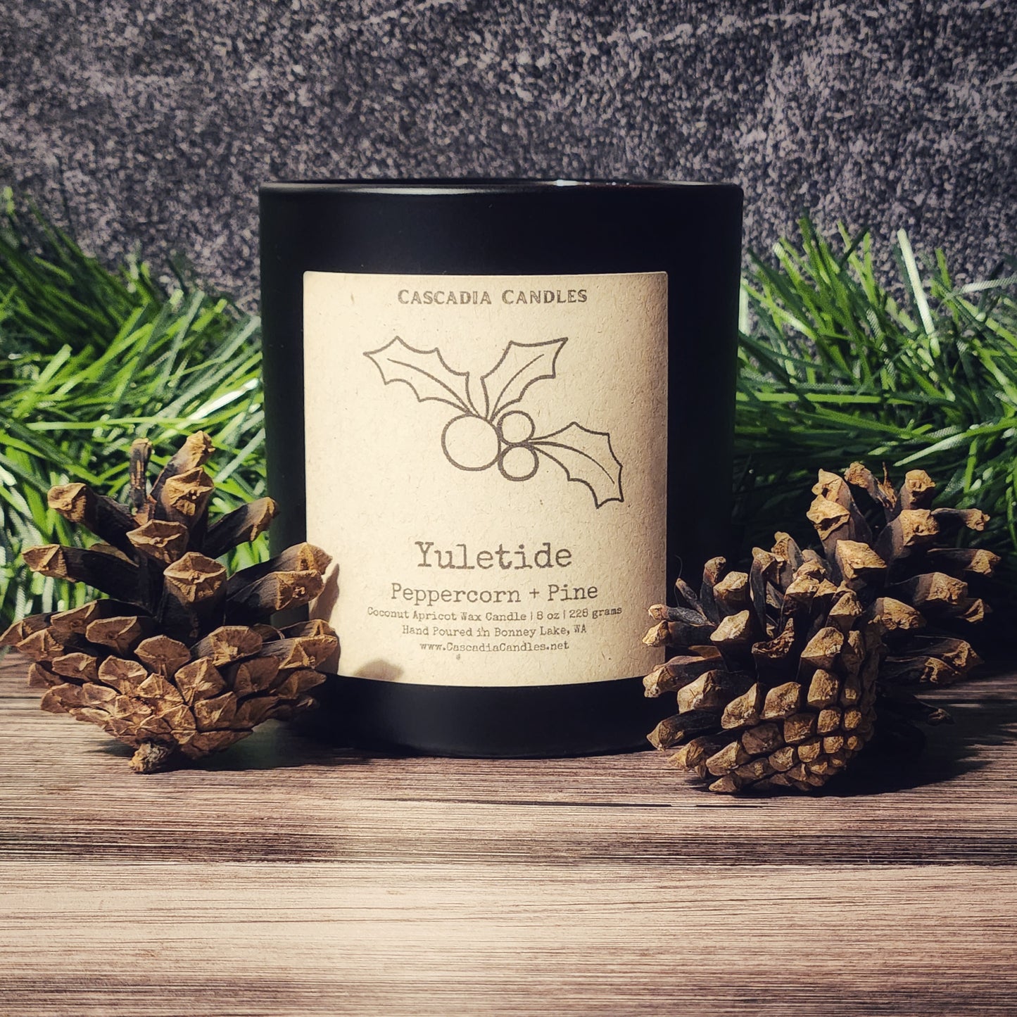 Candle labeled 'Yuletide' with decorative holly design, surrounded by pine cones and greenery.