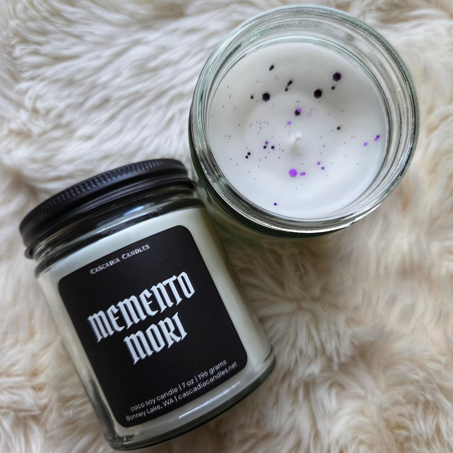 Two jars of candle wax with 'memento mori' text on a fluffy white surface