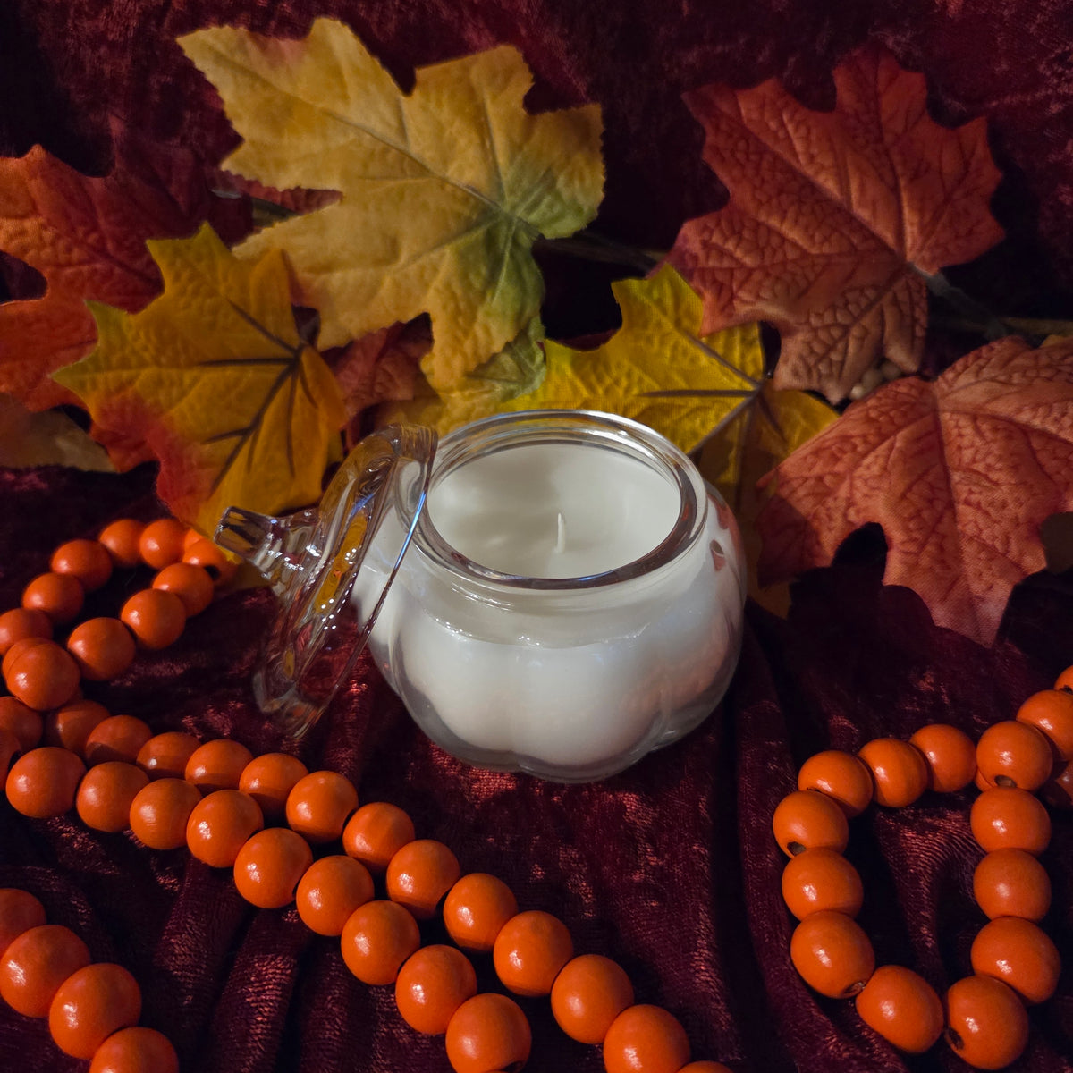 A white candle in a clear glass pumpkin-shaped jar with a glass lid laying on its side, styled on dark red fabric with orange beads and autumn leaves.
