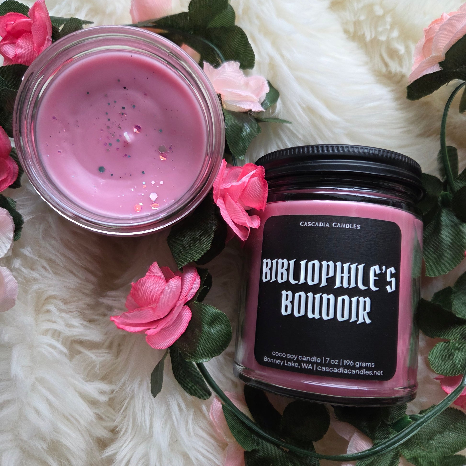 Candle with pink wax and 'Bibliophile's Boudoir' label surrounded by pink flowers on a white surface