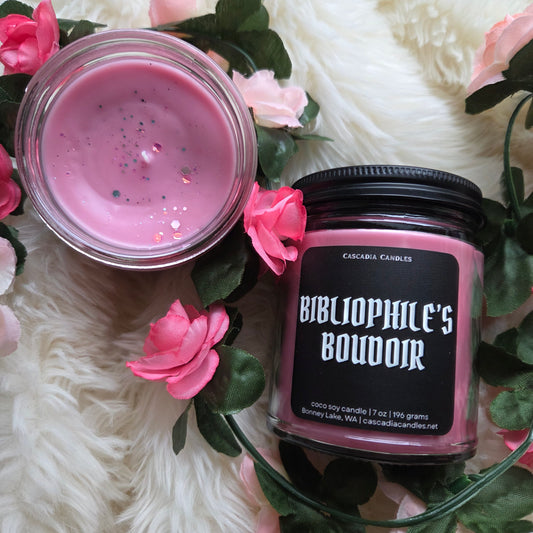 Candle with pink wax and 'Bibliophile's Boudoir' label surrounded by pink flowers on a white surface