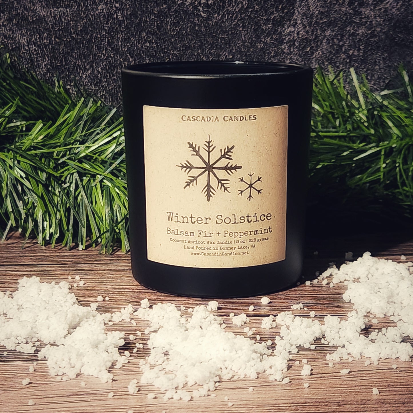 Black candle with a decorative label on a wooden surface with snowflakes and greenery.