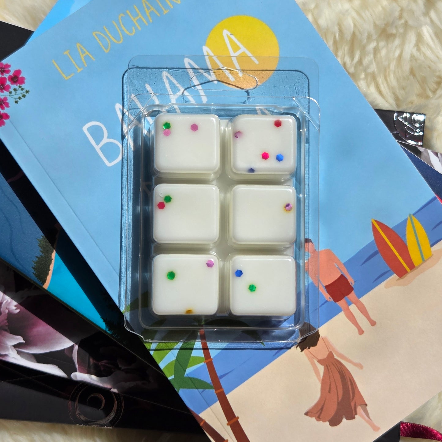 Six white square wax melts with eco-friendly glitter, sealed in a clear clamshell, placed on top of a blue beach-themed book.