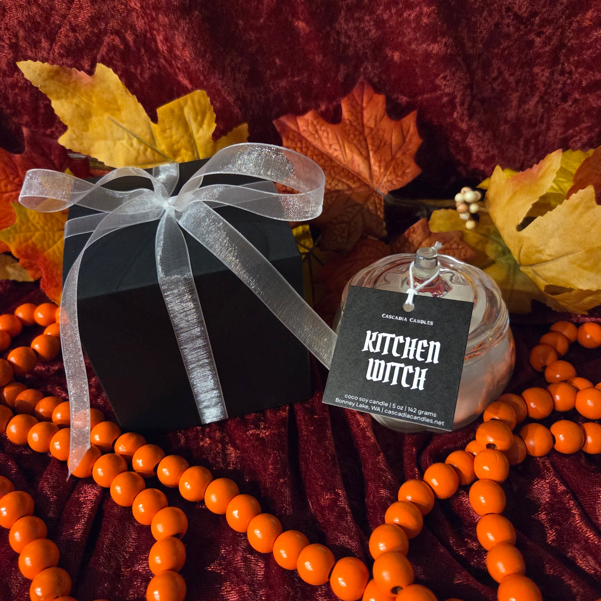 A candle in a clear glass jar with a black tag reading 'KITCHEN WITCH' by Cascadia Candles, styled with a black gift box, white ribbon, orange beads, and autumn leaves on a dark red velvet background.