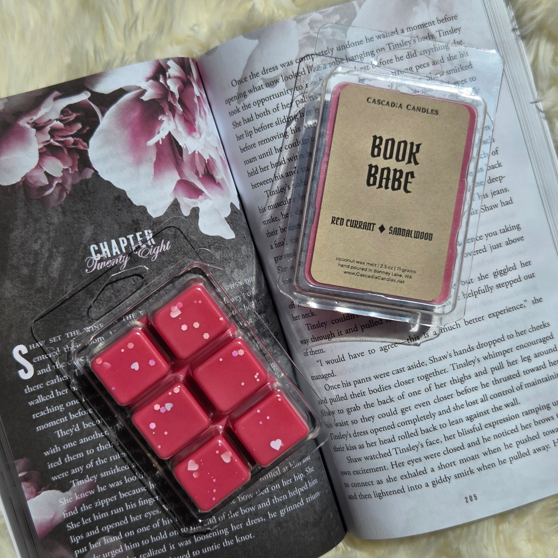 Two wax melts products displayed on an open book: a clamshell of six red wax melts with small white hearts, and a front display of Cascadia Candles Book Babe wax melt that smells like red currant and Sandalwood scent with a Kraft paper label.
