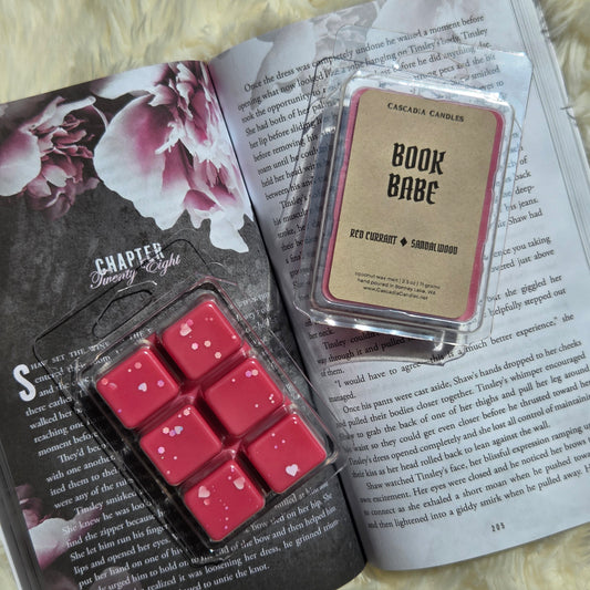 Two wax melts products displayed on an open book: a clamshell of six red wax melts with small white hearts, and a front display of Cascadia Candles Book Babe wax melt that smells like red currant and Sandalwood scent with a Kraft paper label.