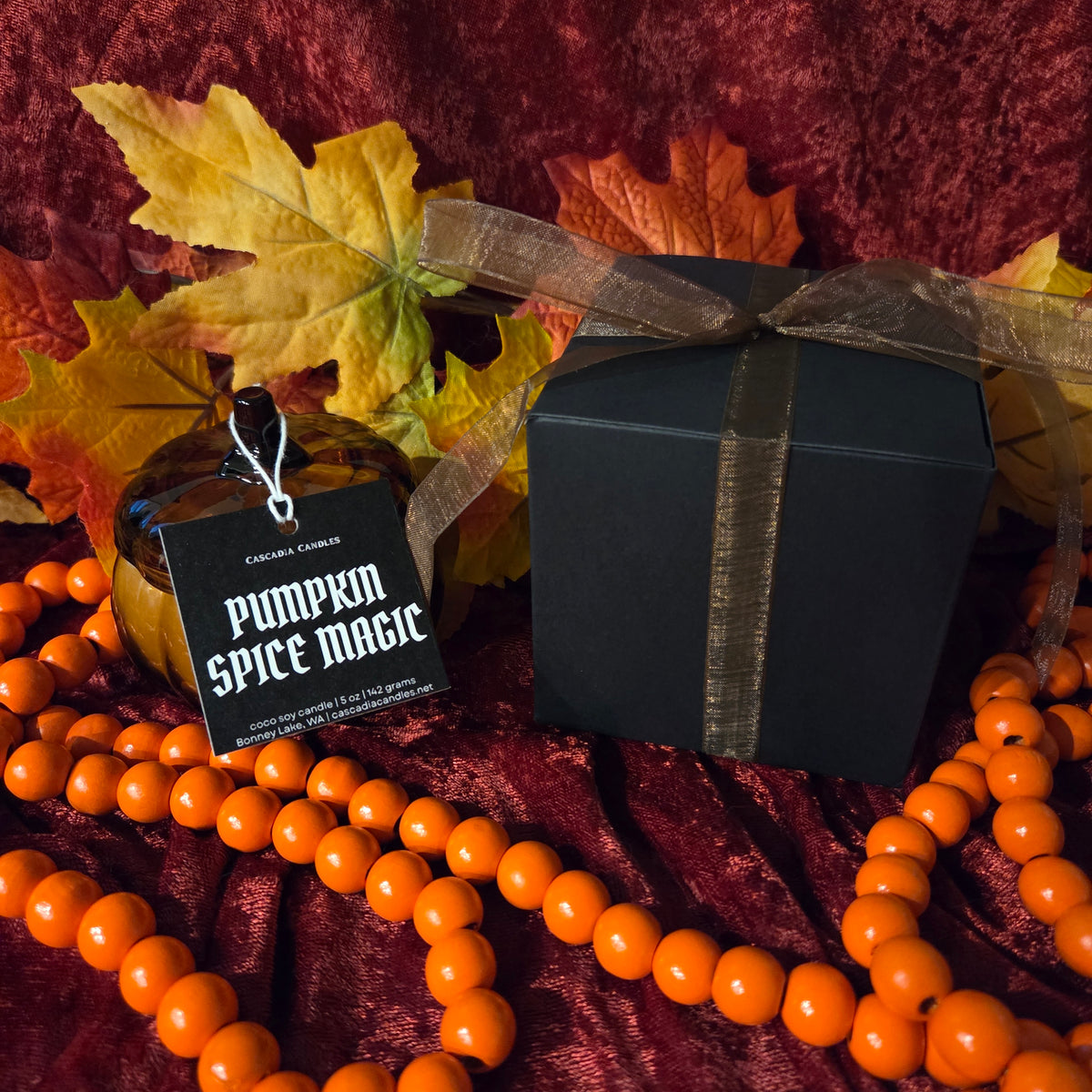 A styled product shot featuring a black gift box tied with a sheer brown ribbon, next to a small glass bottle labeled 'Pumpkin Spice Magic' by Cascadia Candles. The scene is decorated with orange beads, autumn leaves, and a dark red velvet fabric background.