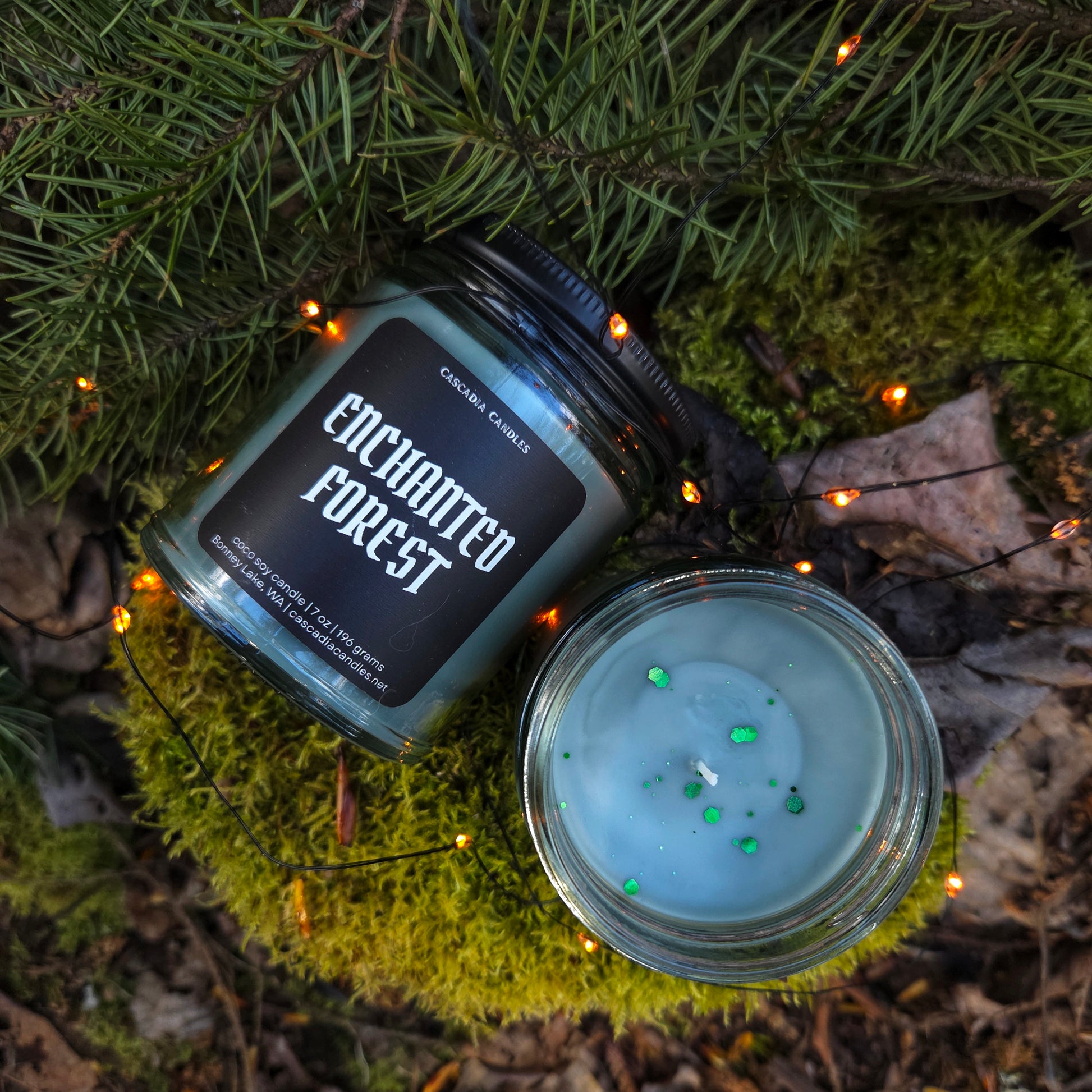 Candle labeled 'Enchanted Forest' on a natural setting with greenery and lights.