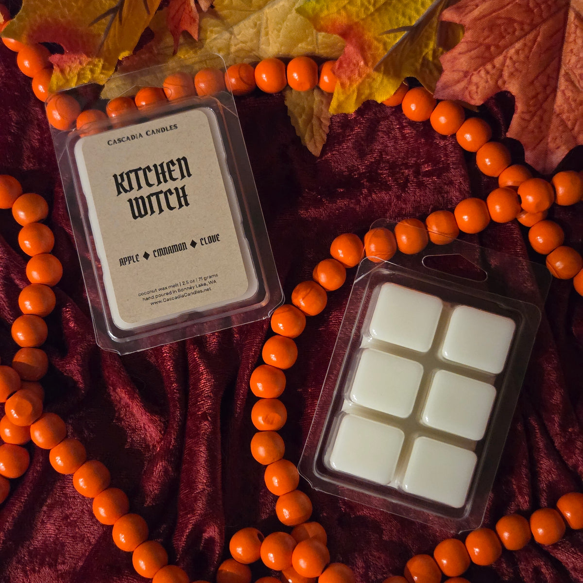 Two packages of Cascadia Candles 'Kitchen Witch' wax melts are arranged on a dark red velvet fabric with orange beaded garland and autumn leaves.