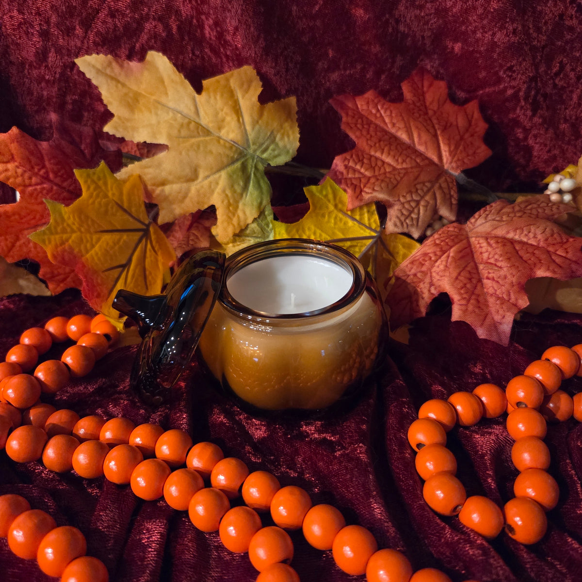 A small amber glass jar pumpkin-shaped candle with a dark glass lid sits on rich burgundy velvet, surrounded by autumn maple leaves and strands of bright orange beads.