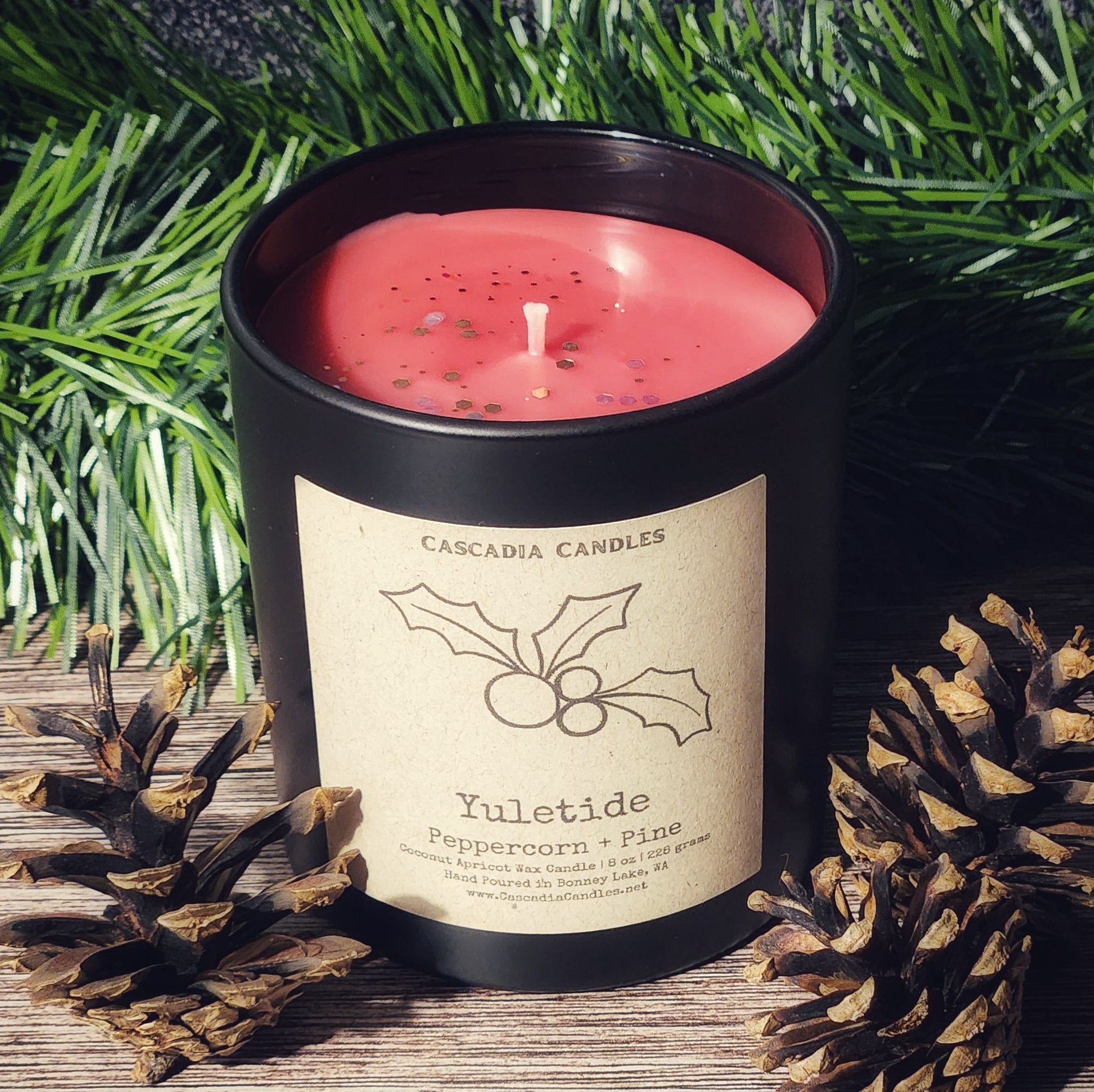 Red candle in a black container with a label on a wooden surface with greenery and pinecones.