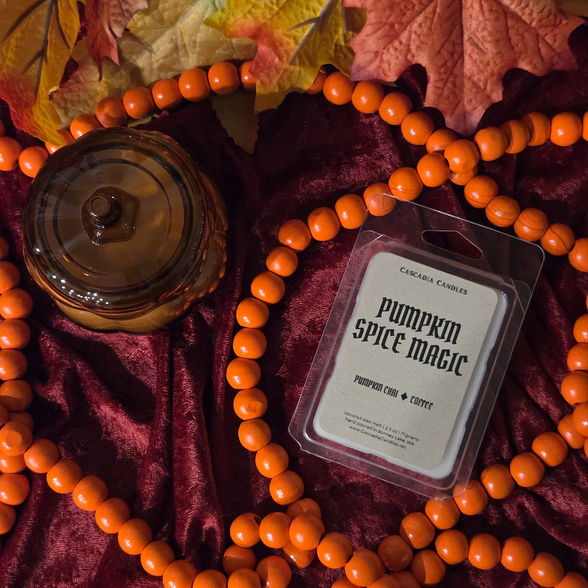 A packaged pumpkin spice candle by Cascadia Candles is displayed on dark burgundy velvet with orange beaded garlands and autumn leaves. A glass pumpkin-shaped jar is also visible in the styled flat-lay arrangement.
